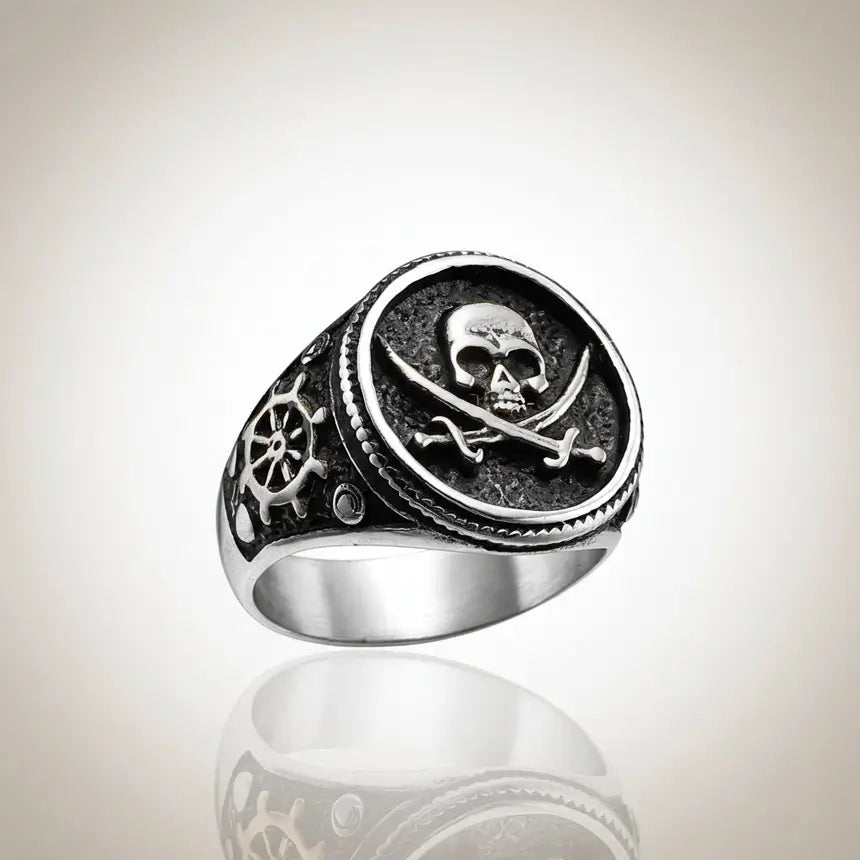 Bague du flibustier maudit en acier 316L finition argentée sur fond clair, design tête de mort et sabres croisés.