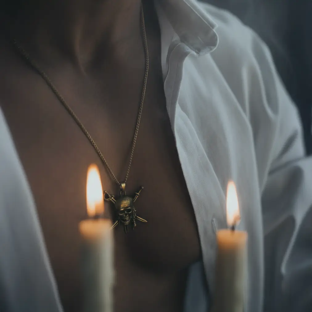 Homme torse nu avec chemise ouverte portant le collier Sabres du Flibustier, reflet doré des bougies sur le pendentif pirate.